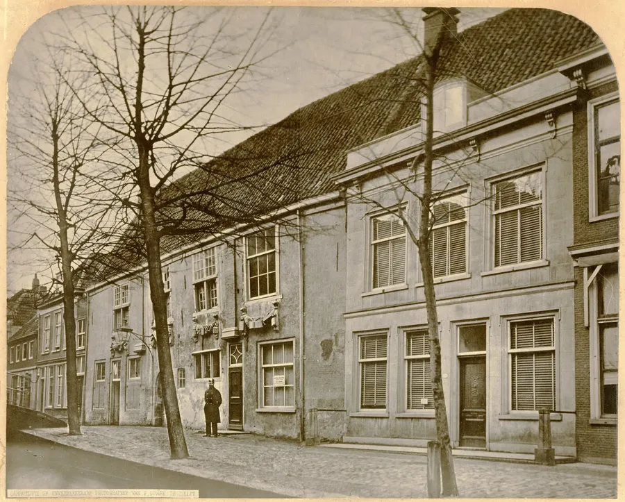 Oude foto van een rij gevels aan de Voldersgracht met in het midden het lage gildehuis met versieringen boven de deur, rechts een gepleisterd herenhuis en kale bomen langs de kade.