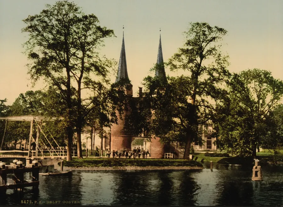 Ingekleurde foto van de Oostpoort met zijn twee spitse torens tussen hoge bomen, aan de overkant van het water, met links een witte ophaalbrug en mensen bij de poort.