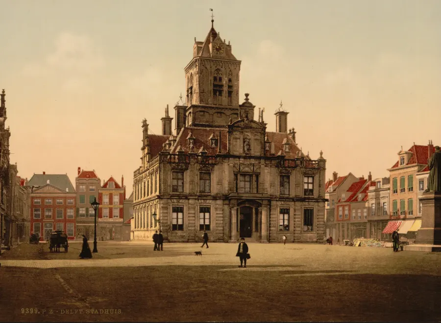 Ingekleurde foto van het stadhuis aan de Markt met de toren erachter, een bijna leeg plein met enkele mensen en een hondje, en rechts aan de rand het standbeeld van Hugo de Groot.