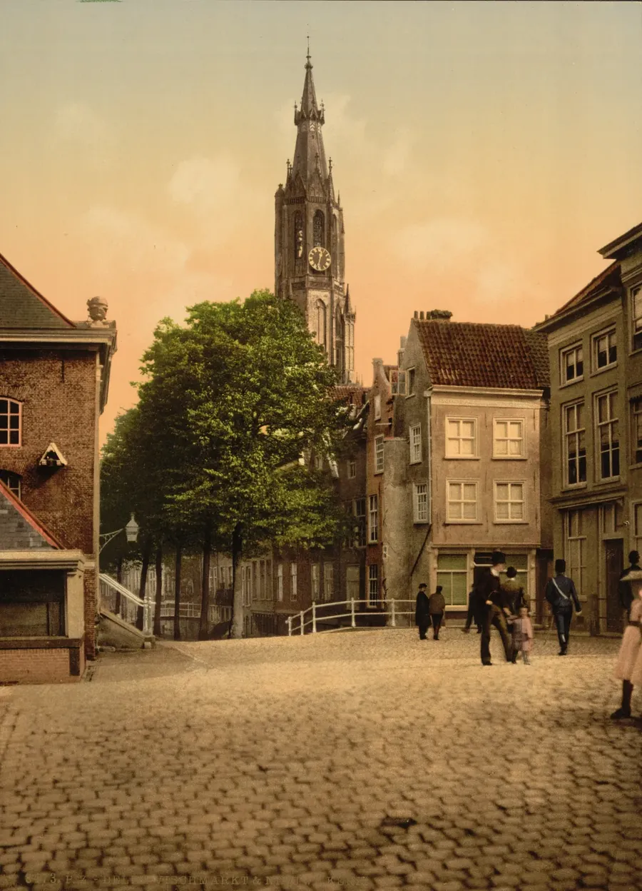 Ingekleurde foto van een geplaveid pleintje met links de Vleeshal, in het midden bomen en een witte brug en daarachter de toren van de Nieuwe Kerk, rechts huizen en een paar wandelaars.
